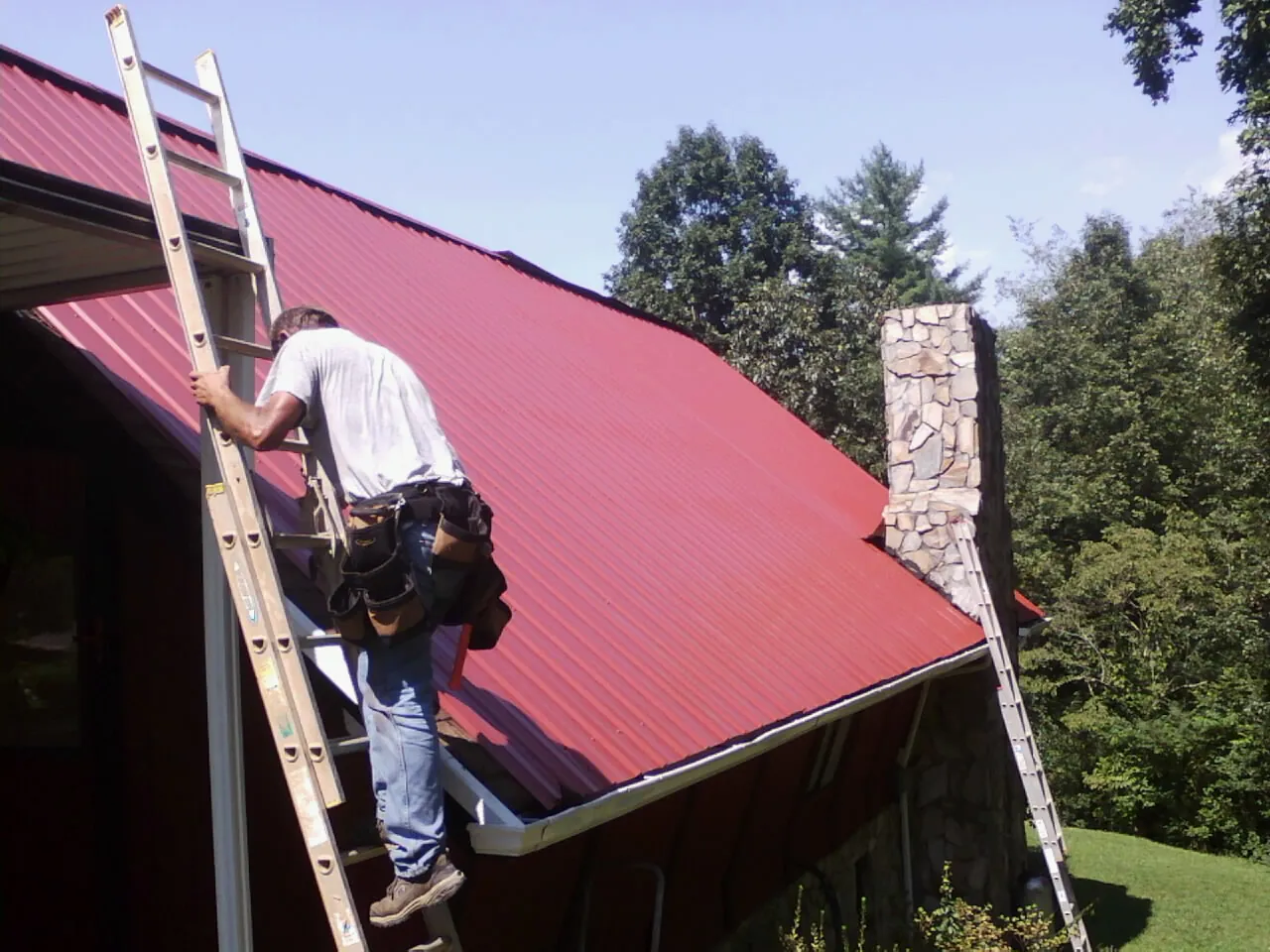Roofer on a ladder beside a red metal roof and stone chimney — Soffit & Fascia Repair by Roofing Brothers in Newbury Park
